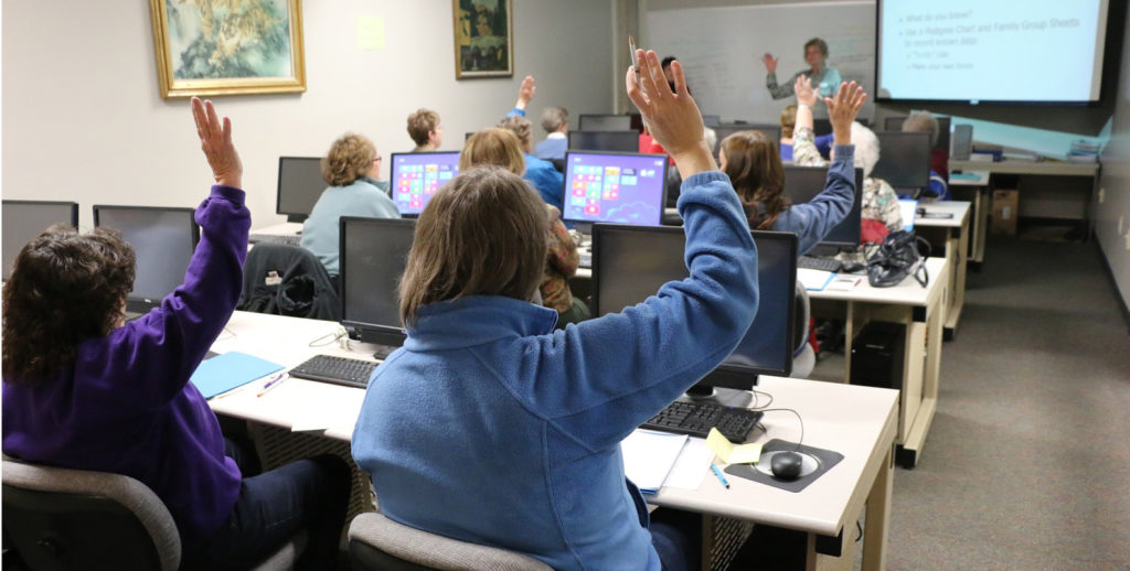 an image of a class room in a computer lab, raising their hands to address the teach.er